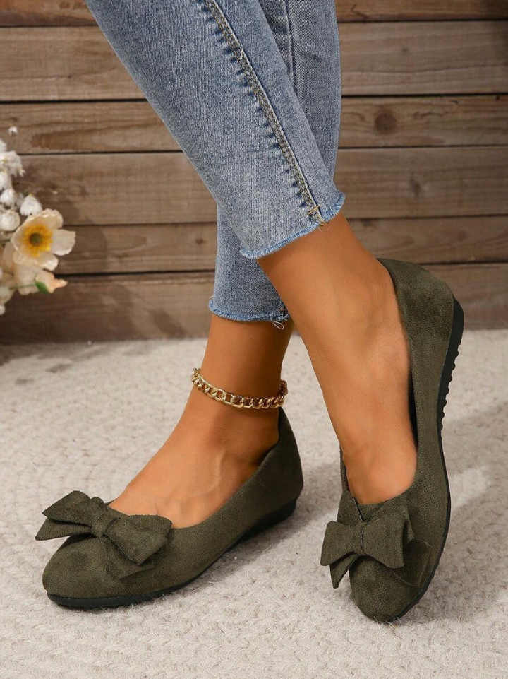 Green flat shoes with bow details worn with blue jeans on a wooden floor.