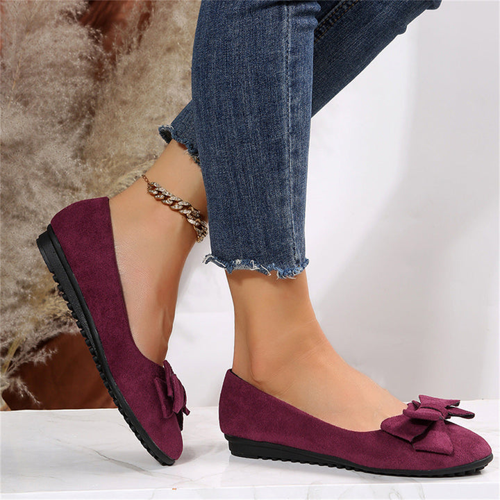 Purple flats with bow details worn with blue jeans on a neutral background