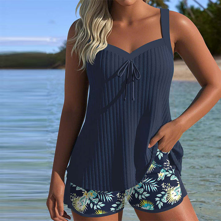 Female model wearing a striped tankini swimsuit with beach shorts on a plain background.