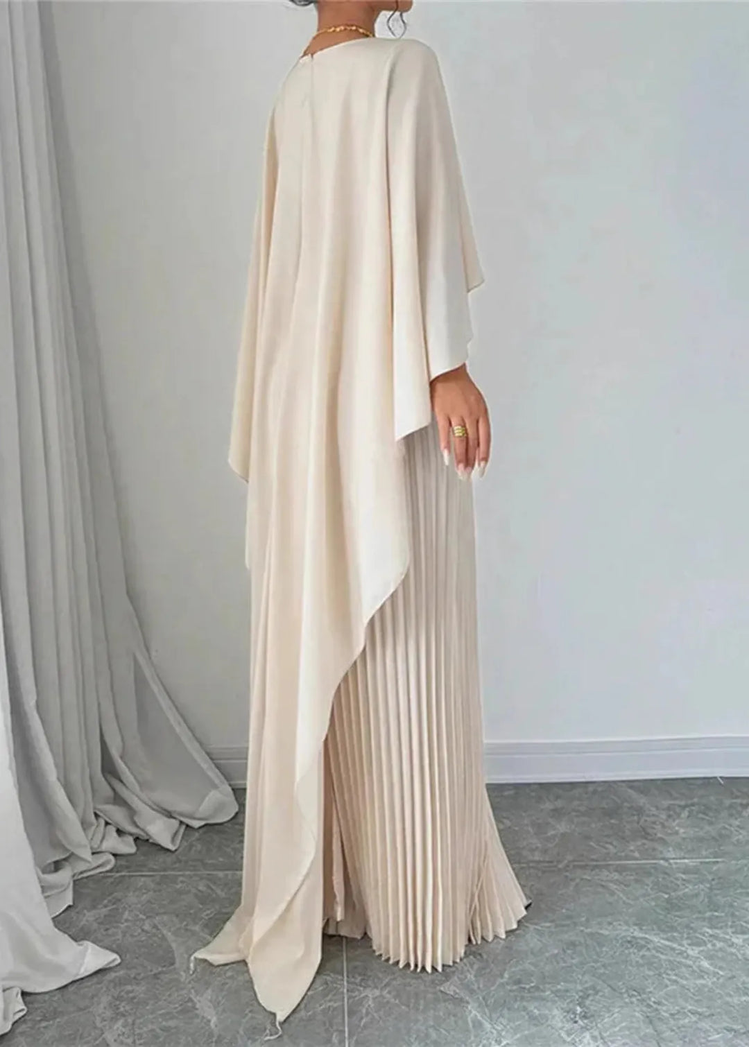 Female model in a flowing pleated maxi dress with cape detail on a plain background.