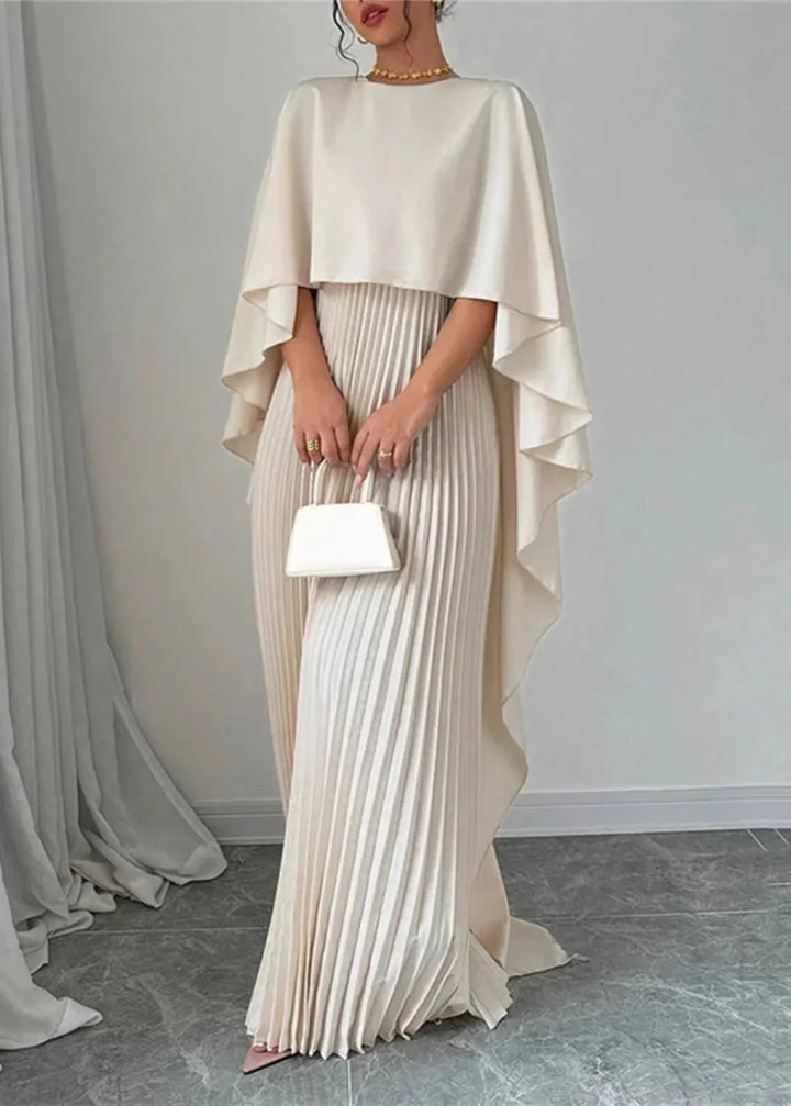 Woman showcasing an elegant cape maxi dress with soft pleats on a plain background.