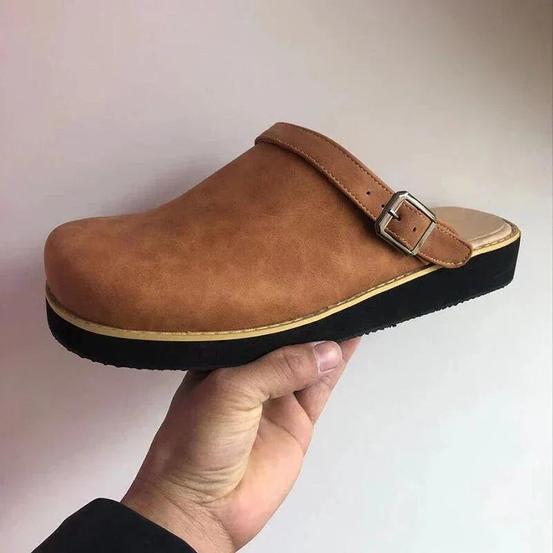 Brown clog shoe with a strap held by a hand against a plain background