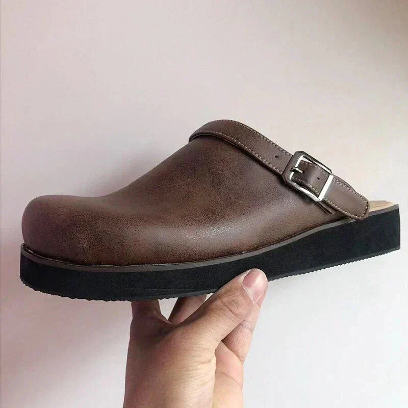 Brown clog shoe with a black sole held by a hand against a plain background