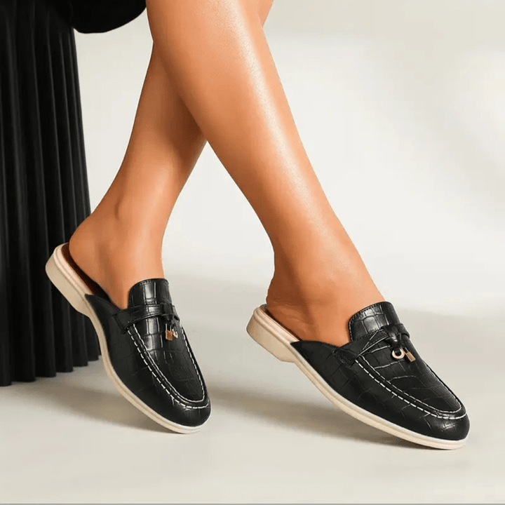 Foot wearing stylish mule indoors, plain beige background shot.