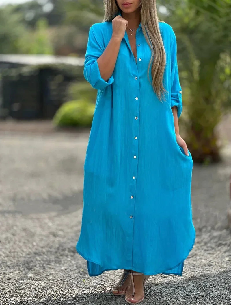 Woman styled in a long relaxed shirt dress with roll-up sleeves on plain background.