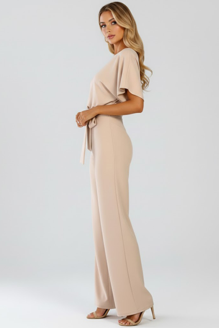 Model wearing a full-length jumpsuit with round neckline, waist tie, and relaxed sleeves on a plain background.