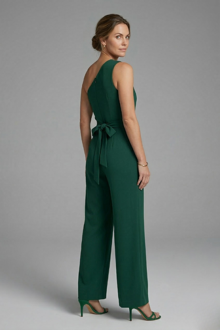 Woman in a wide leg sleeveless jumpsuit with asymmetric neckline and belt detail on a plain background.