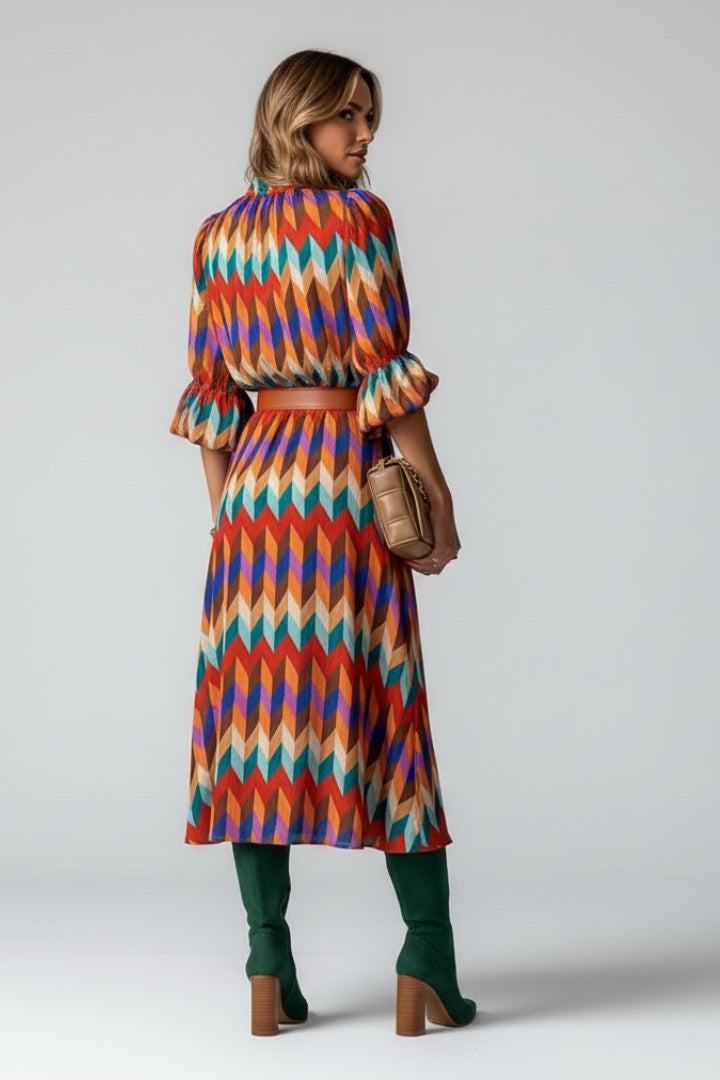 Female model standing in multicolour printed dress against plain gray background, back angle