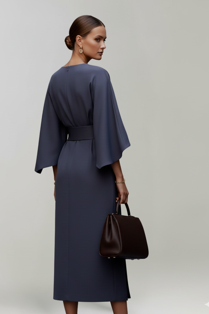 Female model wearing elegant belted dress holding a bag, back pose, gray background