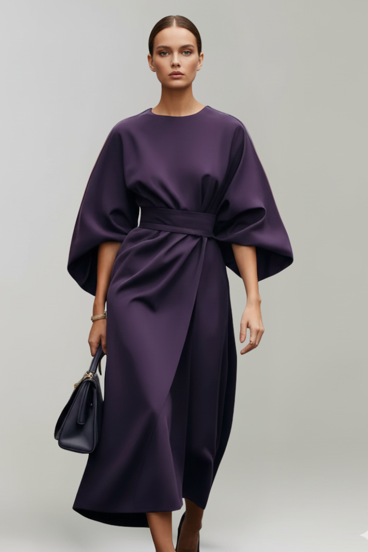 Woman standing in belted waist dress on plain gray background.