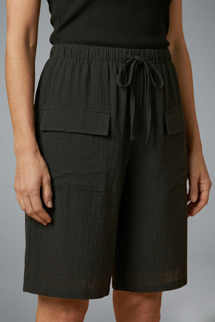 Woman showing front pocket detail on shorts, plain gray background.