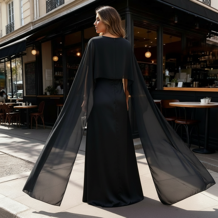 Woman posing in a satin-style evening gown with cape sleeves on a plain background.