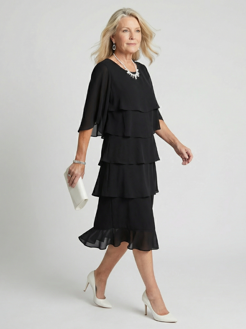 Woman posing in a flowing midi dress with layered skirt detailing on a plain background.