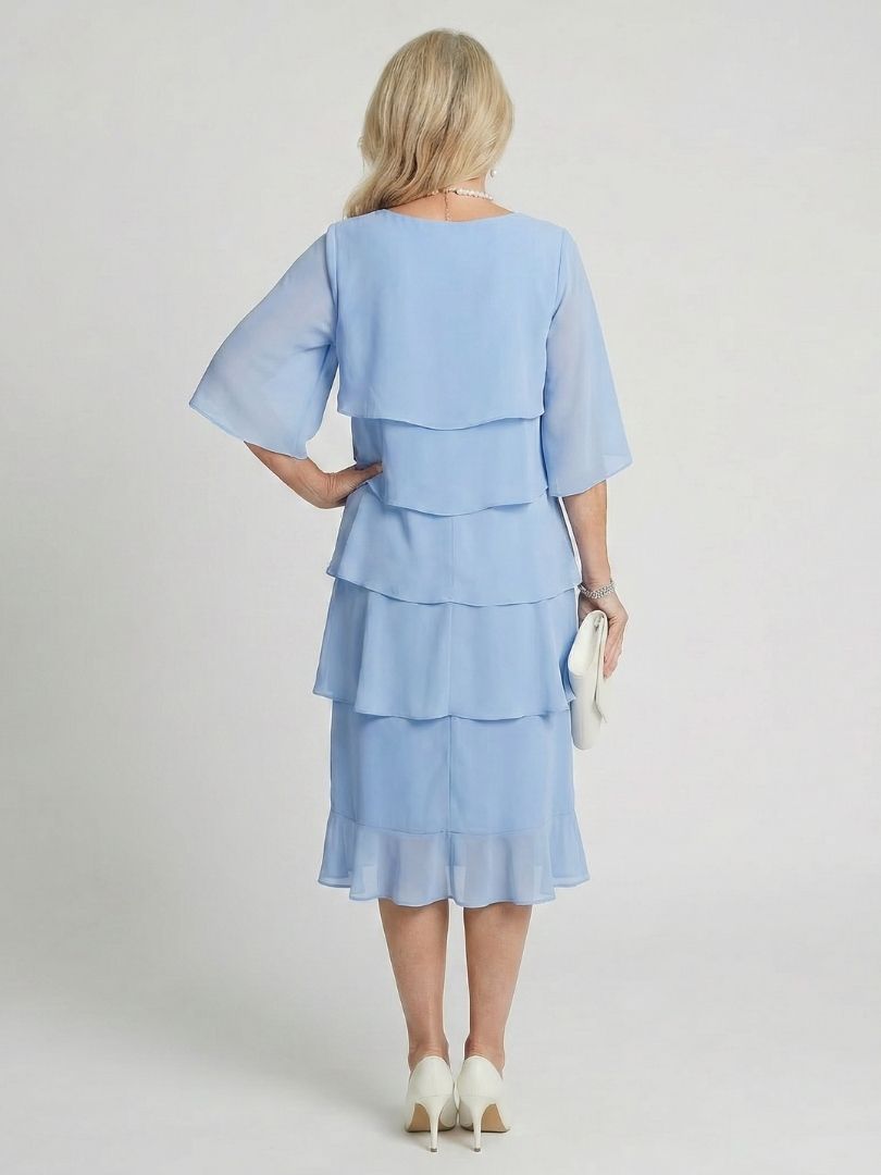 Woman standing in a layered midi dress with soft drape and flow.