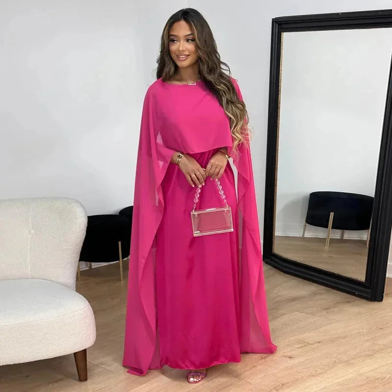 Woman in a long cape dress styled for wedding guest occasions on a plain background.