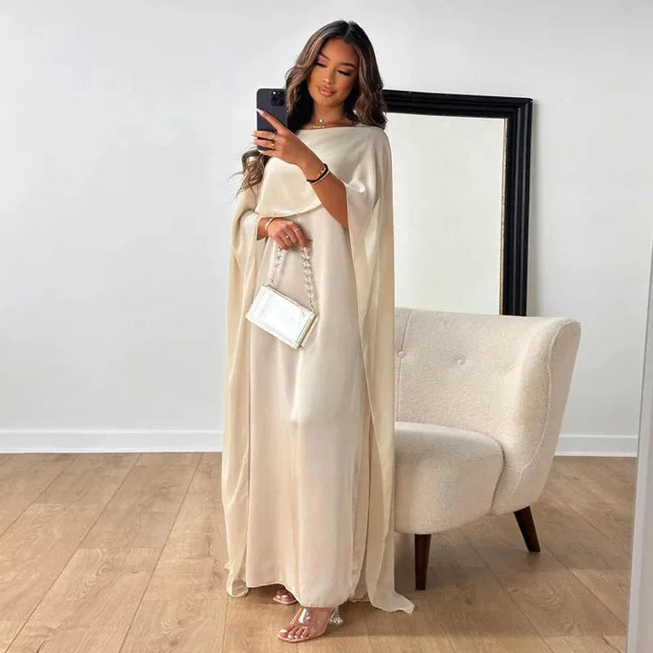 Female model wearing a cape maxi dress with a flowing silhouette against a plain background.
