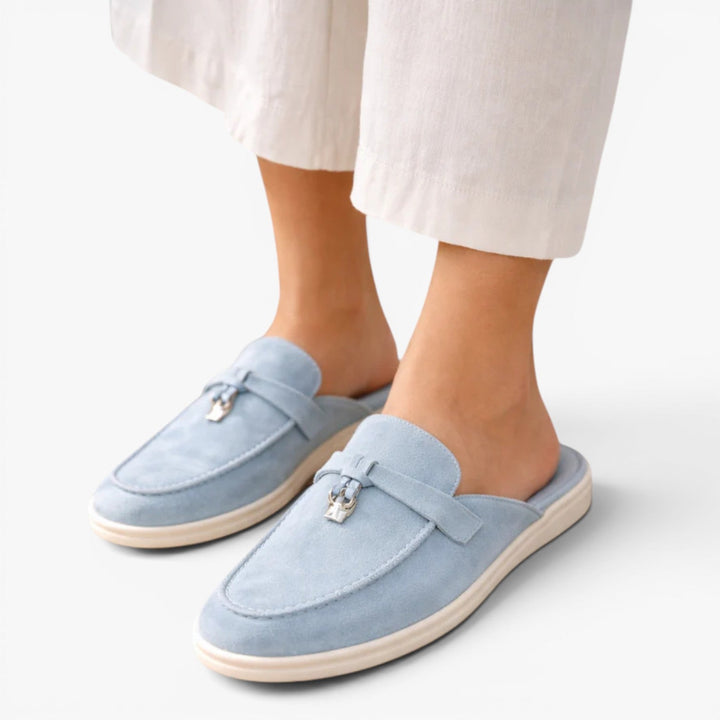  Loafer-inspired mule shoes with decorative hardware on a plain background.