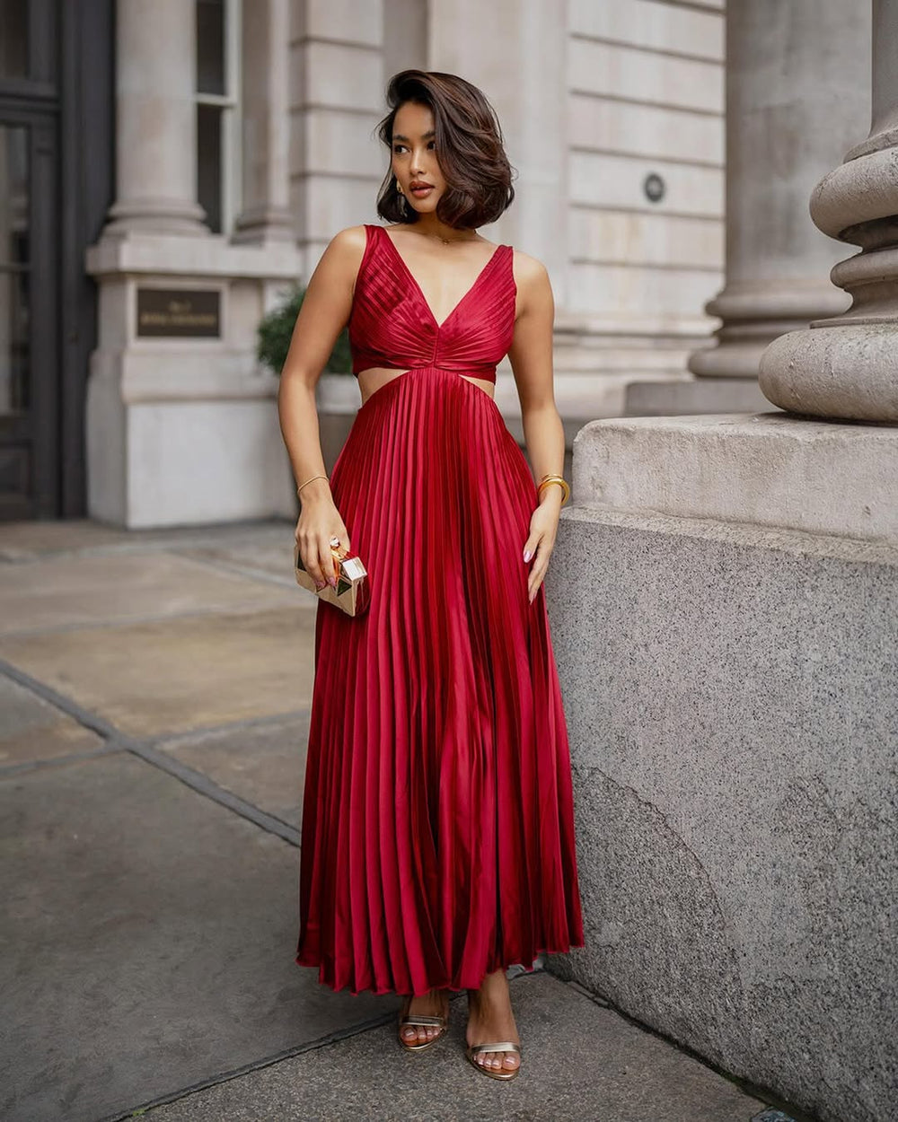 Woman showcasing a flowing pleated midi dress with a waist cutout detail on a plain background.